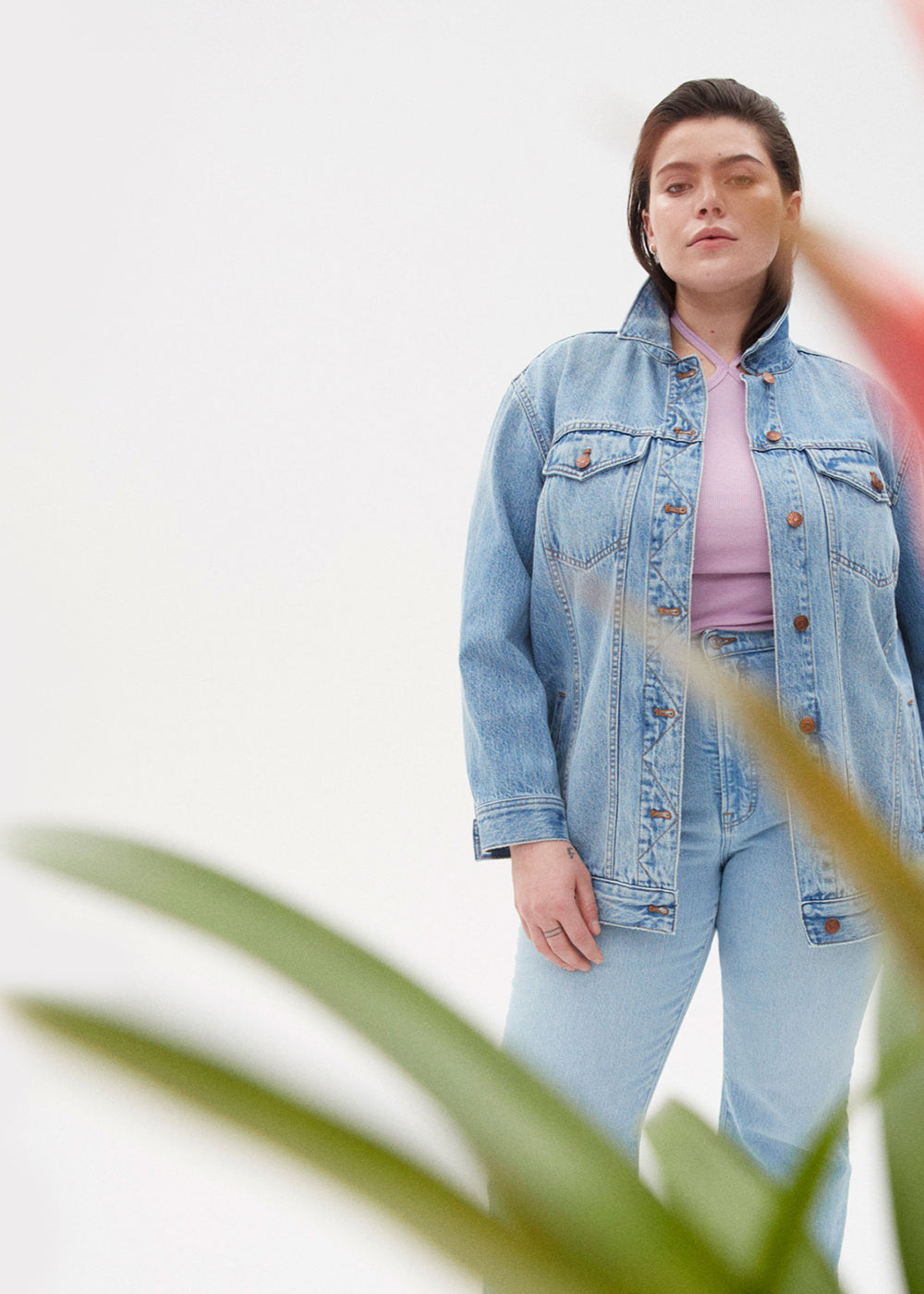 Individual posing behind a bromeliad wearing a Madewell denim jacket, Madewell jeans, and a Madewell lavender halter tank top