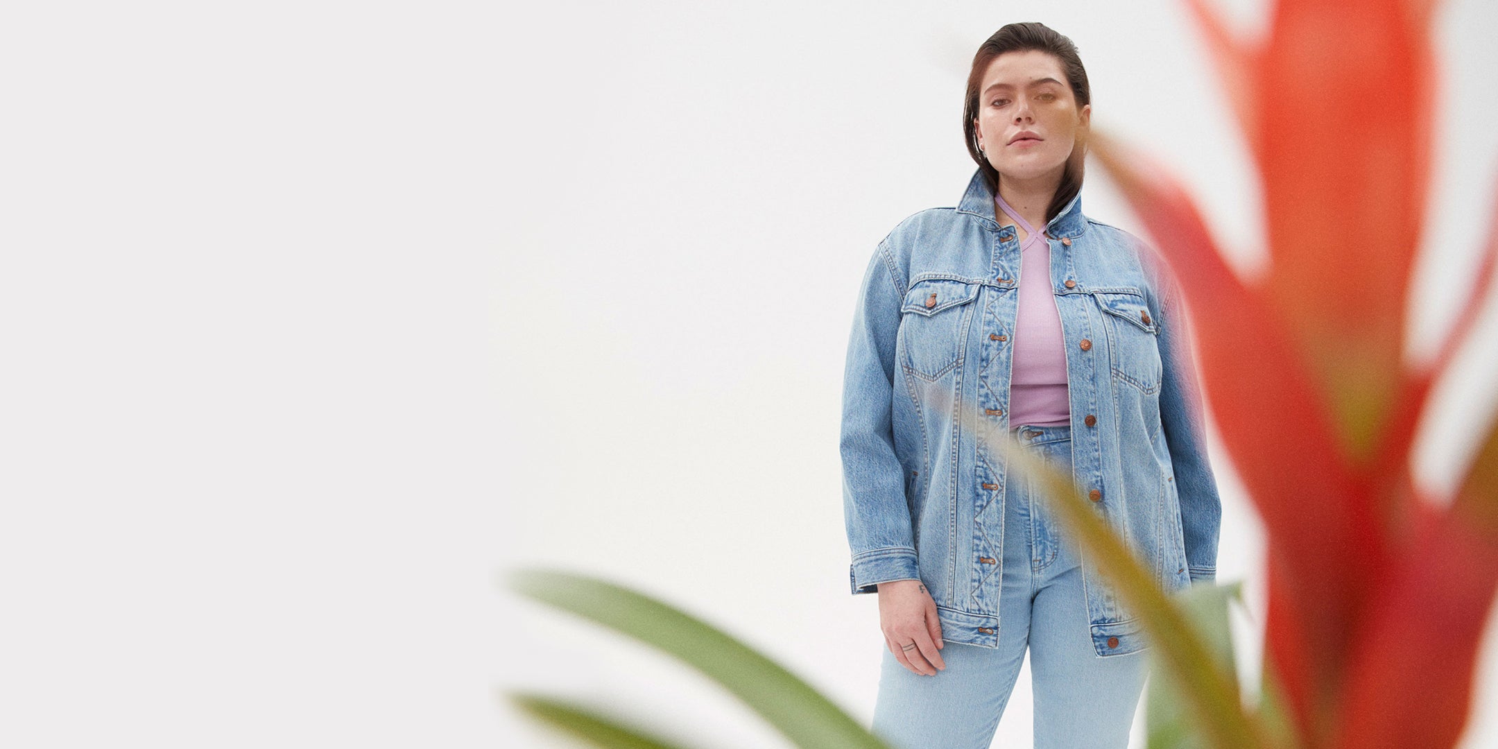 Individual posing behind a bromeliad wearing a Madewell denim jacket, Madewell jeans, and a Madewell lavender halter tank top